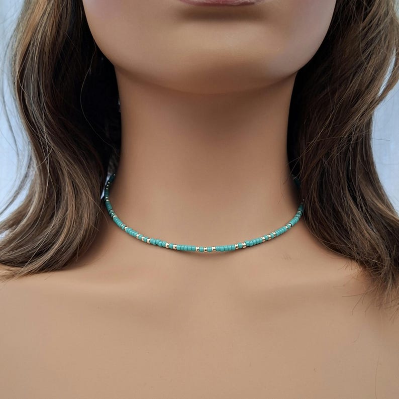 May include: A delicate choker necklace featuring a repeating pattern of small, round turquoise-colored beads interspersed with tiny, metallic silver-toned spacer beads, creating a simple and uniform circular design around the neck.
