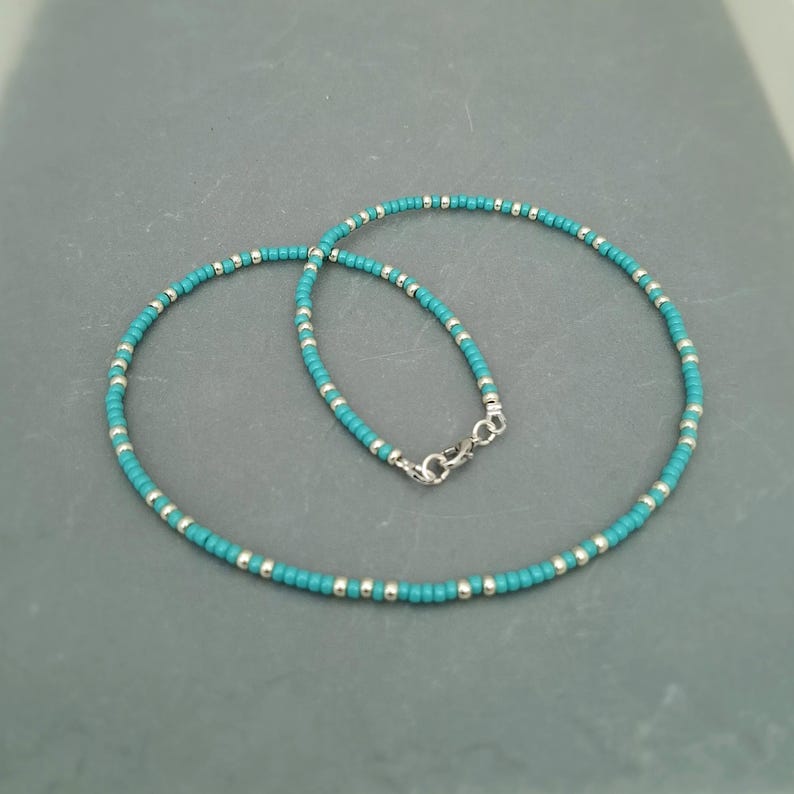 May include: A beaded necklace featuring a repeating pattern of small, round turquoise-colored beads and metallic silver-toned spacer beads. The necklace is secured with a silver-toned lobster claw clasp and is arranged in a circular shape.