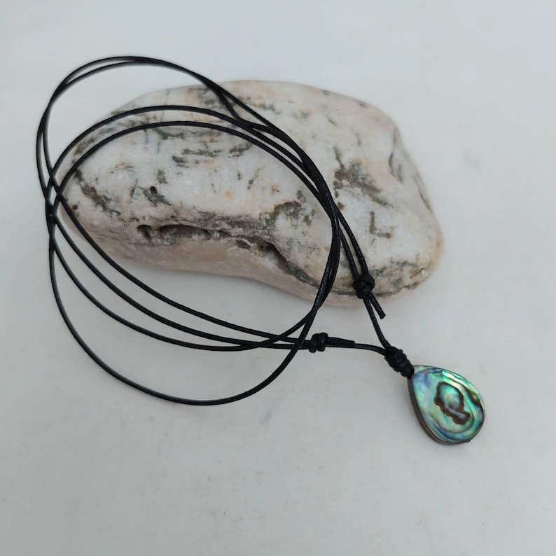 May include: A black cord necklace with a teardrop-shaped abalone shell pendant. The shell has iridescent green, blue, and gold colors.