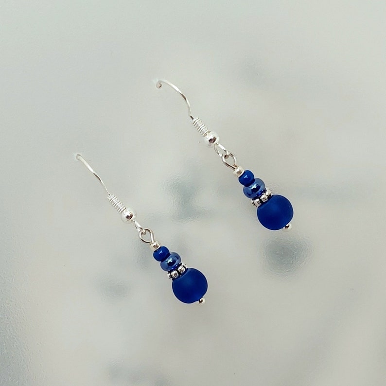 May include: A pair of silver dangle earrings with blue glass beads. The earrings have a simple design and are perfect for everyday wear.