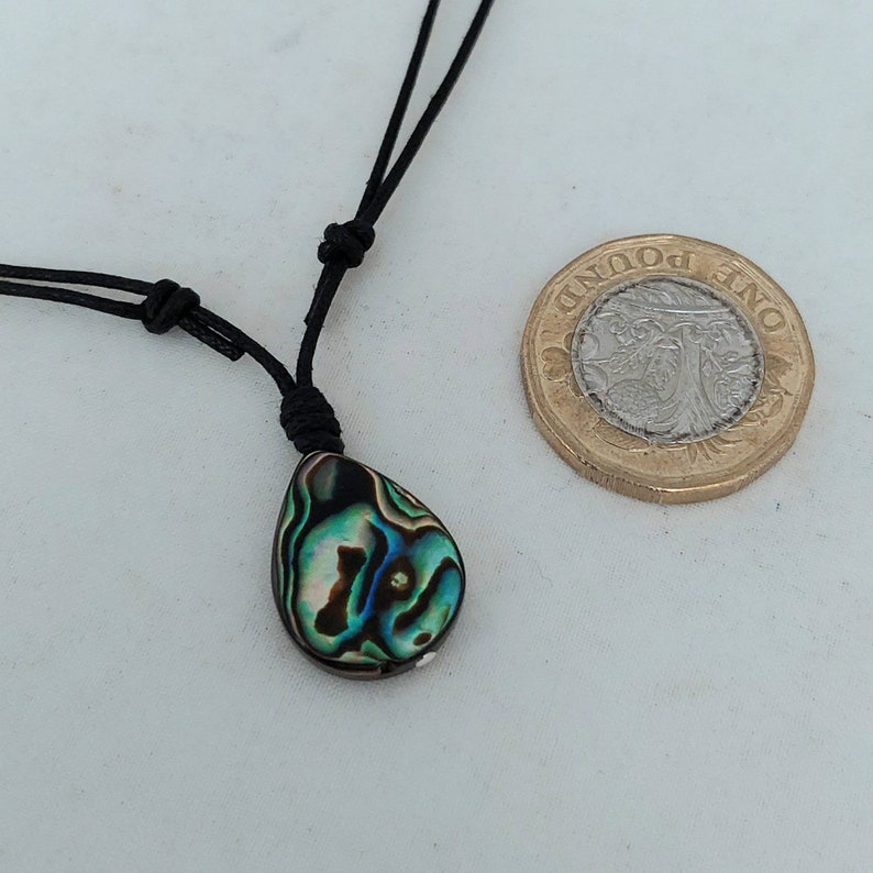 May include: A teardrop-shaped abalone shell pendant on a black cord necklace. The pendant has iridescent green, blue, and black colors. The necklace is shown next to a British one pound coin for size reference.