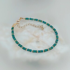 May include: A delicate bracelet made with small teal and gold beads. The bracelet has a gold clasp and extender chain.