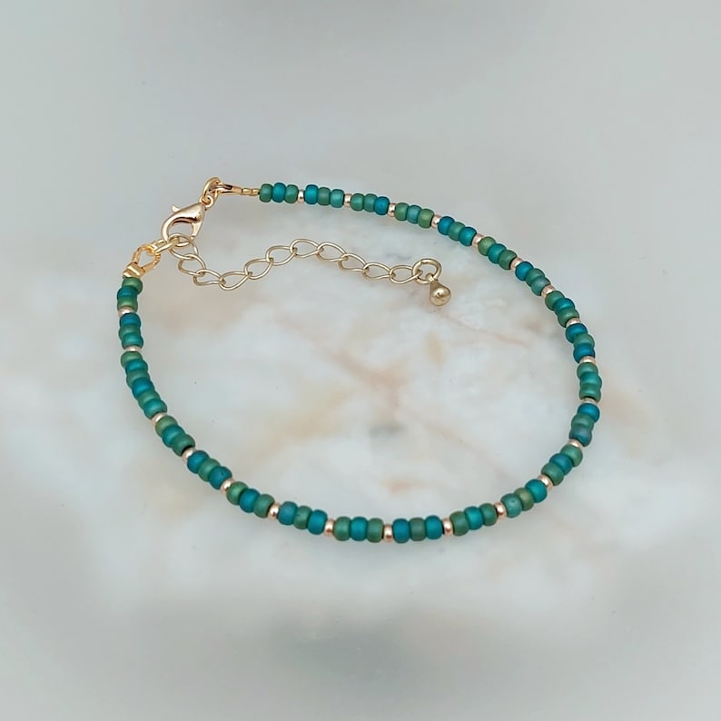 May include: A delicate bracelet made with small teal and gold beads. The bracelet has a gold clasp and extender chain.