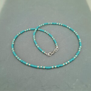 May include: A beaded necklace featuring a repeating pattern of small turquoise blue seed beads and metallic silver spacer beads. The necklace is secured with a silver-toned lobster claw clasp and a small jump ring, creating a circular shape.