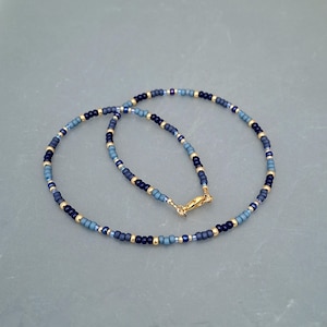 May include: A beaded necklace featuring a repeating pattern of small round beads in shades of navy blue, slate blue, and metallic gold. The necklace is secured with a gold-toned lobster claw clasp, creating a circular shape with a consistent design.