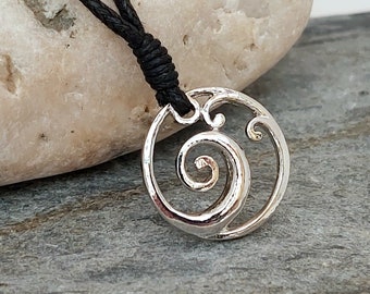 May include: Silver necklace with a circular pendant featuring a stylized wave design. The pendant is attached to a black cord.