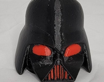 Puede incluir: Una figura de plástico negro de Darth Vader con ojos rojos. La figura es un modelo impreso en 3D del personaje de Star Wars.