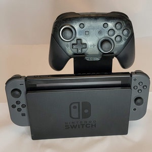 May include: A black Nintendo Switch console with a black controller attached to the top. The console is sitting on a white surface. The console has the text "NINTENDO SWITCH" printed on the front.