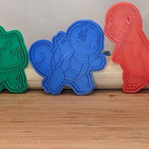 Puede incluir: Tres cortadores de galletas con forma de personajes de Pokémon: un Bulbasaur verde, un Squirtle azul y un Charmander rojo.