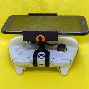 May include: A white Xbox controller with a black and orange phone holder attached to it. A black smartphone is in the holder.