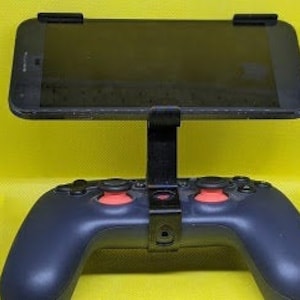 Peut inclure: Un smartphone noir est monté sur un support noir, qui est fixé à une manette de jeu vidéo noire avec des boutons rouges. La manette est sur une surface jaune.