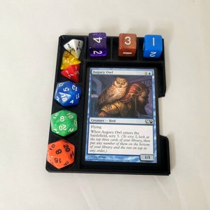 May include: A black plastic game tray with a Magic: The Gathering card titled "Augury Owl" and several colorful dice. The card text reads "When Augury Owl enters the battlefield, scry 3. (To scry 3, look at the top three cards of your library, then put any number of them on the bottom of your library and the rest on top in any order.)"