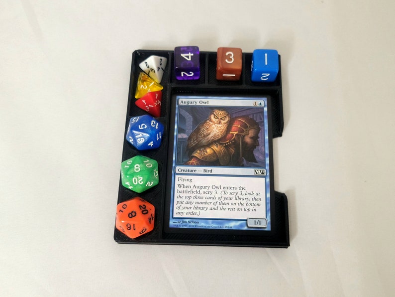 May include: A black plastic game tray with a Magic: The Gathering card titled "Augury Owl" and several colorful dice. The card text reads "When Augury Owl enters the battlefield, scry 3. (To scry 3, look at the top three cards of your library, then put any number of them on the bottom of your library and the rest on top in any order.)"
