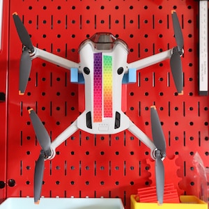 May include: A white drone with black propellers and a rainbow patterned sticker on the top. The drone is hanging on a red pegboard.