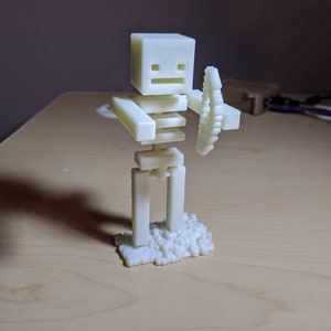 Puede incluir: Una figura blanca de un esqueleto sosteniendo una espada, impresa en 3D. El esqueleto est&aacute; de pie sobre una base de peque&ntilde;os bloques.