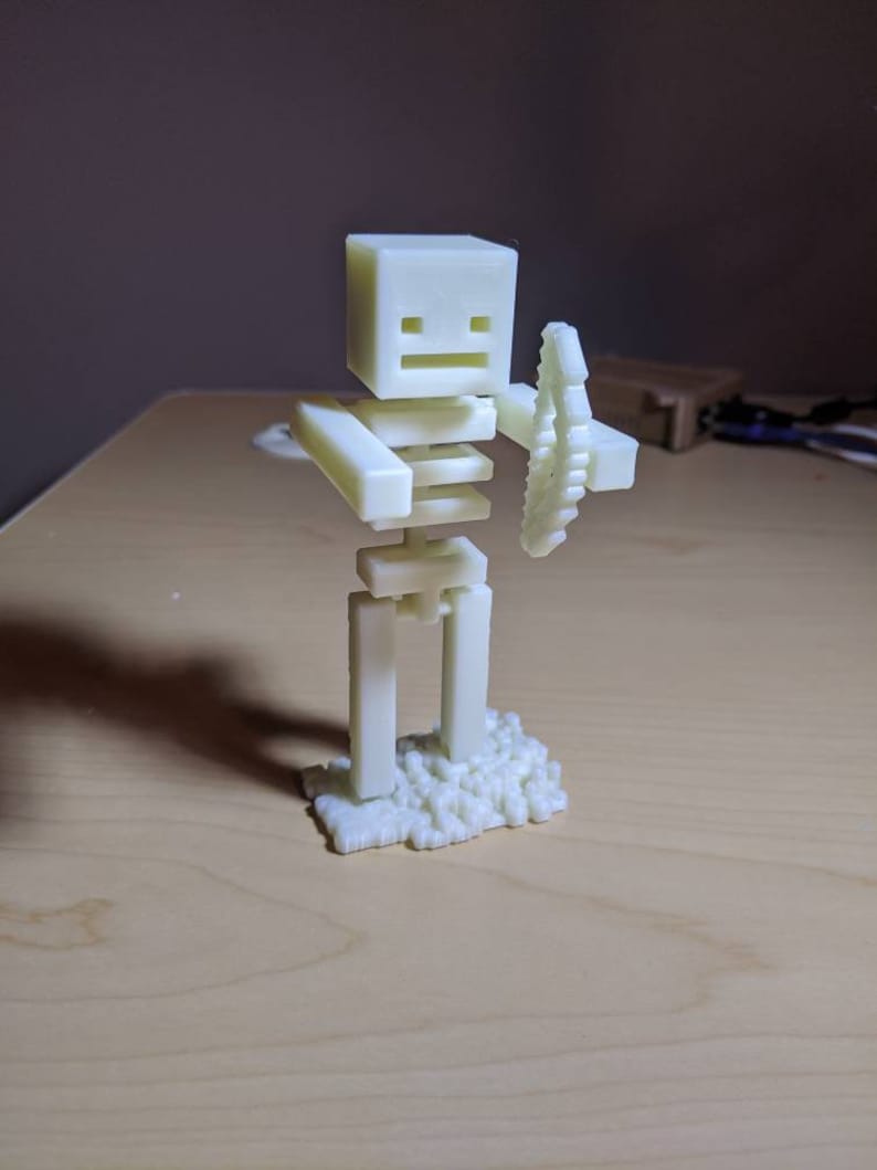 Puede incluir: Una figura blanca de un esqueleto sosteniendo una espada, impresa en 3D. El esqueleto est&aacute; de pie sobre una base de peque&ntilde;os bloques.