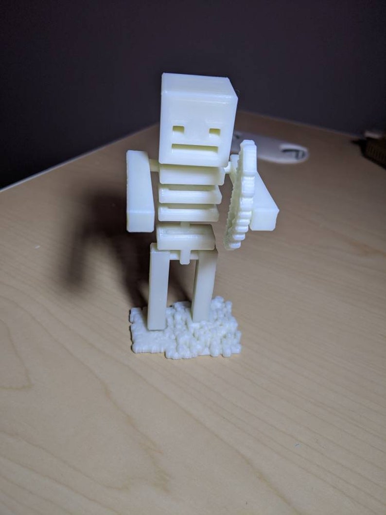 Puede incluir: Figurita blanca impresa en 3D de un esqueleto sosteniendo un objeto circular. El esqueleto est&aacute; de pie sobre una base de peque&ntilde;os cubos blancos.
