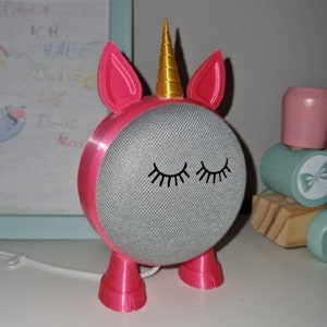 May include: A pink unicorn-shaped stand for a Google Home smart speaker. The stand has a gold horn and black eyelashes. The speaker is gray and has a fabric mesh covering.