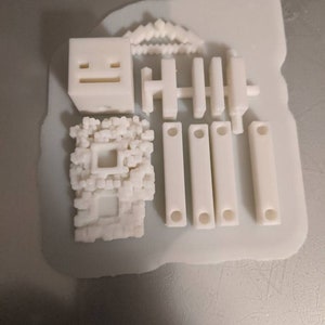 Puede incluir: Un conjunto de bloques de construcci&oacute;n de pl&aacute;stico blanco impresos en 3D, que incluye un cubo con una cara, una valla y un peque&ntilde;o edificio con una ventana.