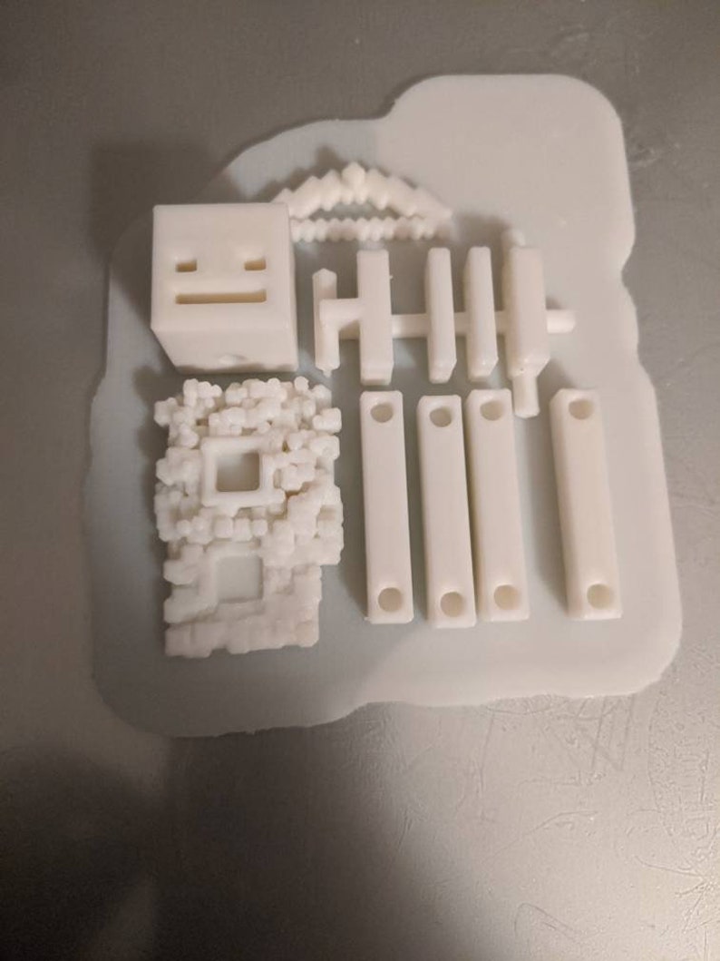 Puede incluir: Un conjunto de bloques de construcci&oacute;n de pl&aacute;stico blanco impresos en 3D, que incluye un cubo con una cara, una valla y un peque&ntilde;o edificio con una ventana.