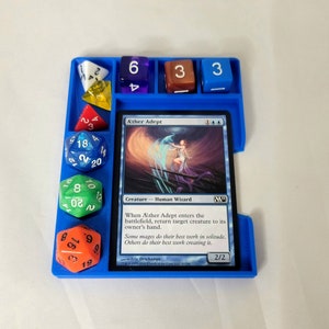 May include: A blue plastic dice tray with a Magic: The Gathering card titled "&AElig;ther Adept" and several dice. The card text reads "When &AElig;ther Adept enters the battlefield, return target creature to its owner's hand. Some mages do their best work in solitude. Others do their best work creating it."  The card has a 2/2 power and toughness.