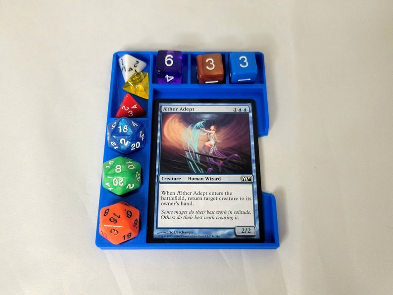 May include: A blue plastic dice tray with a Magic: The Gathering card titled "&AElig;ther Adept" and several dice. The card text reads "When &AElig;ther Adept enters the battlefield, return target creature to its owner's hand. Some mages do their best work in solitude. Others do their best work creating it."  The card has a 2/2 power and toughness.