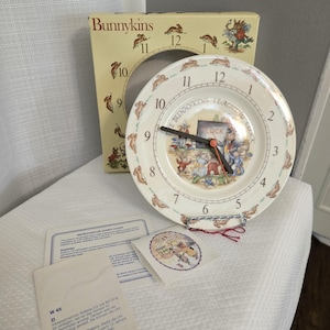 May include: A Bunnykins ceramic clock with a cream-coloured face, black hands, and an orange second hand. The clock features a colourful illustration of rabbits and the words "Bunnykins Teaching Clock". The clock is approximately 20 cm in diameter.