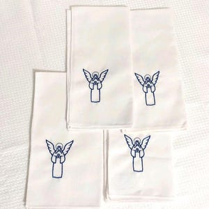 Może przedstawiać: Pięć białych ręcznik&oacute;w z niebieskim haftowanym wzorem anioła. Ręczniki są złożone i ułożone na białej, teksturowanej powierzchni. Anioł ma rozłożone skrzydła i złożone dłonie.