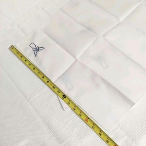 Może przedstawiać: Biała kwadratowa chusteczka z niebieskim haftowanym wzorem w rogu. Ż&oacute;łta taśma miernicza jest umieszczona po przekątnej na chusteczce, pokazując pomiar około 48 cm.