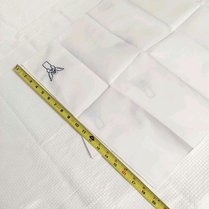 Może przedstawiać: Biała kwadratowa chusteczka z niebieskim haftowanym wzorem w rogu. Ż&oacute;łta taśma miernicza jest umieszczona po przekątnej na chusteczce, pokazując pomiar około 48 cm.