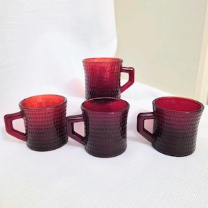 Puede incluir: Un juego de cuatro tazas de vidrio de color rojo intenso con un patrón texturizado de puntos en el exterior. Cada taza tiene un asa resistente y una forma cilíndrica. El vidrio es translúcido, permitiendo que la luz atraviese el color rojo.