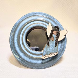 May include: A round, ceramic mirror with a light blue and gray finish. A ceramic angel with white wings and a blue dress is attached to the frame. The mirror is a decorative item.