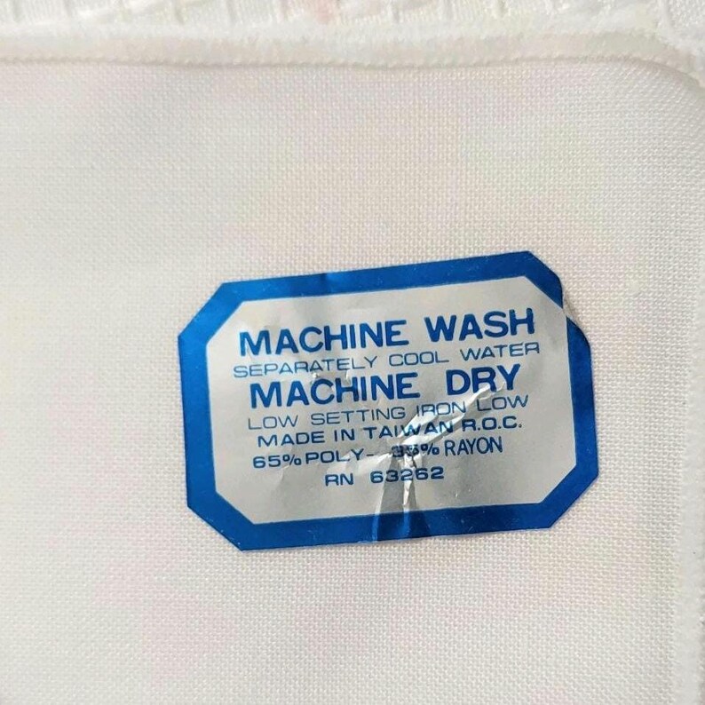 Może przedstawiać: Biała tkanina z niebiesko-białą etykietą. Etykieta zawiera napisy "MACHINE WASH" i "MACHINE DRY" z instrukcjami pielęgnacji. Tkanina wykonana jest w 65% z poliestru i rayonu.