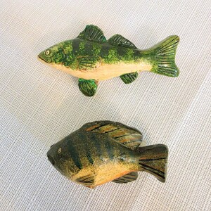 May include: Two carved wooden fish figurines. The top fish features a green and tan striped pattern with detailed fins. The bottom fish is darker, with a deep olive and brown mottled finish, rounded body shape, and defined fin markings.