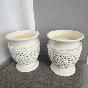 Puede incluir: Dos candelabros de cerámica blanca con intrincados diseños recortados. Los candelabros tienen una base ancha y redonda y un cuerpo cilíndrico con un borde festoneado.