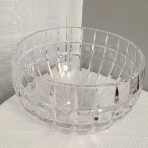 May include: A clear, colorless glass bowl featuring a geometric, grid-like cut pattern across its entire exterior surface. The bowl has a rounded shape with a wide opening and a thick, faceted rim, creating a reflective and textured appearance.