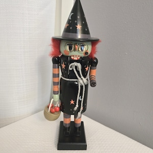 May include: A Halloween-themed witch nutcracker. The witch has a green face, red hair, and wears a black dress with orange accents. The witch is holding a basket with red apples and wears a black hat with stars and a crescent moon.