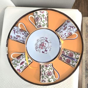 May include: A set of six small teacups and a saucer arranged in a circular box. The cups and saucer are light blue with a floral design in pink and green. The box has an orange interior and a brown rim.