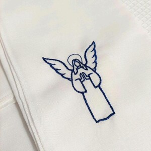 Może przedstawiać: Biała tkanina z szczeg&oacute;łowym niebieskim haftem przedstawiającym anioła. Anioł jest przedstawiony z rozpostartymi skrzydłami, aureolą i dłońmi złożonymi do modlitwy. Tkanina ma subtelną fakturę, a haft jest starannie wykonany.