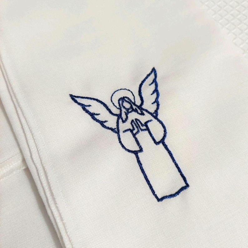 Może przedstawiać: Biała tkanina z szczeg&oacute;łowym niebieskim haftem przedstawiającym anioła. Anioł jest przedstawiony z rozpostartymi skrzydłami, aureolą i dłońmi złożonymi do modlitwy. Tkanina ma subtelną fakturę, a haft jest starannie wykonany.