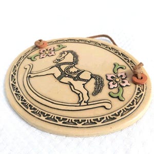 May include: A round, cream-colored ceramic wall plaque featuring a black-outlined rocking horse illustration in the center. The design includes pink and green floral accents, a decorative border, and two brown leather cords attached for hanging.