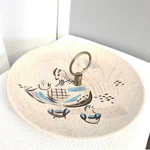 Puede incluir: Bandeja de servir vintage de dos niveles con superficie crema moteada. Cuenta con un asa dorada y un diseño caprichoso en azul y marrón, que representa aves y barcos estilizados. Perfecta para servir aperitivos o postres.