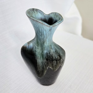 Puede incluir: Un jarrón de cerámica con un borde acampanado en forma de corazón y un cuello estrecho. El esmalte transiciona desde un azul hielo claro en la parte superior hasta un negro profundo en la base, con vetas verdes que mezclan ambos colores.