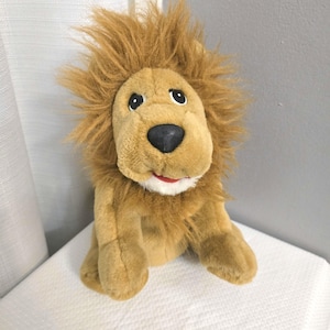 May include: A plush lion toy with a tan body, a brown mane, and a black nose. The lion has a smiling red mouth and black eyes. The toy is sitting on a white surface.