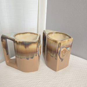 May include: Two ceramic mugs with a unique square design and a handle. The mugs have a tan base with brown and blue glaze. Each mug features a decorative ram's head on the side.
