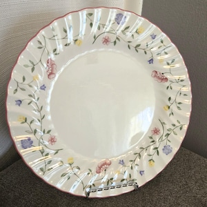 May include: A white ceramic dinner plate with a floral pattern in shades of pink, yellow, blue, and green. The plate has a scalloped edge and a pink rim.