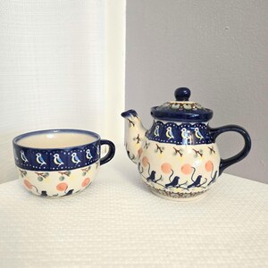 May include: A ceramic teapot and teacup set. The teapot has a dark blue lid and handle, with a white body decorated with blue birds, pink circles, and green branches. The teacup has a dark blue rim and handle, with a matching design.