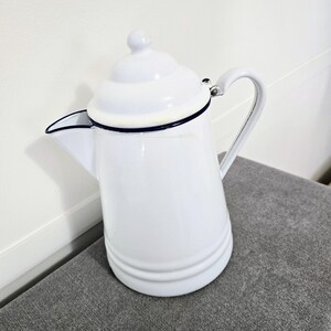 May include: A white enamel coffee pot with a lid and handle. The pot has a black trim around the top and spout. The pot is sitting on a gray surface.