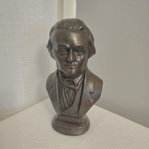 May include: A bronze-coloured bust sculpture of a man in a suit with a bow tie. The sculpture is mounted on a pedestal. The bust is a detailed representation of a historical figure.