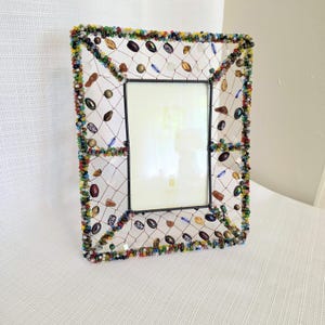 May include: A rectangular picture frame featuring a central glass pane surrounded by a wire mesh border. The outer edge and the mesh are decorated with a variety of small, multi-colored glass beads and larger, assorted oval-shaped beads in various colors.